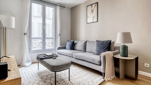 Un salon moderne avec un canapé gris, une petite table d’appoint, une lampe et une fenêtre avec des rideaux.