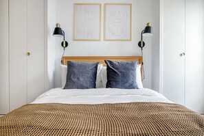 A neatly made bed with a wooden headboard, two pillows, and a textured brown blanket. There are two wall-mounted lamps on either side of the bed. - Blueground | Miromesnil, elev, nr Monceau Parc (Paris)