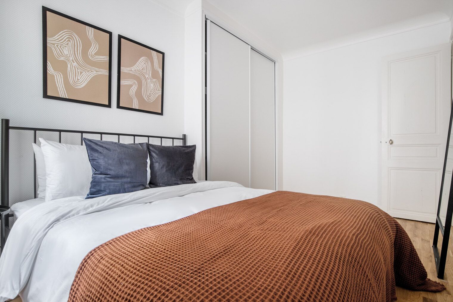 A neatly made bed with white and brown bedding, a black metal headboard, and two abstract wall art pieces.