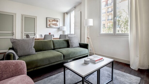 A modern living room with a green sofa, a coffee table, and a view of buildings through large windows. - Blueground | Arc de Triomphe, nr shopping (Paris)