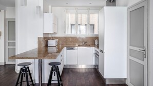 A modern kitchen with white cabinetry, a granite countertop, a built-in dishwasher, and a stainless steel refrigerator. There are two bar stools and a hanging pendant light. - Blueground | Arc de Triomphe, nr shopping (Paris)
