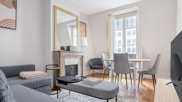 Un salon moderne avec un canapé gris, une cheminée, une table à manger avec des chaises et une grande fenêtre donnant sur des immeubles.