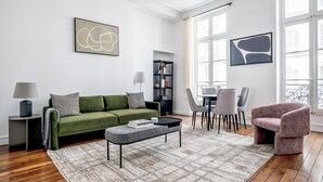 A modern living room with a green sofa, a grey ottoman, and a dining area with chairs and a table.