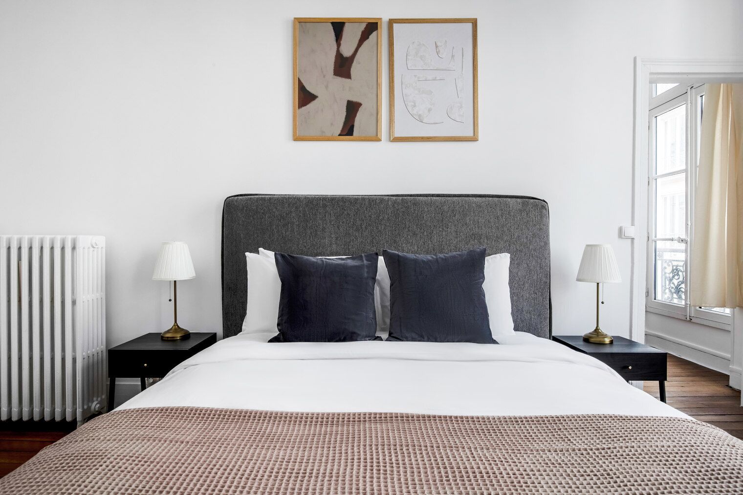 A modern bedroom with a large bed, two bedside tables with lamps, a radiator, and abstract wall art.