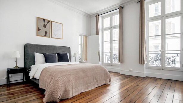 A spacious bedroom with a large bed, wooden flooring, and large windows offering a view of the city.