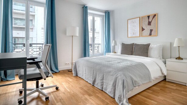 Een moderne slaapkamer met een bed, bureau en stoel, grote ramen met gordijnen en uitzicht op de stad.