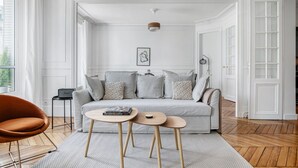 Un salon spacieux avec un canapé gris clair, deux petites tables d’appoint en bois et un fauteuil en cuir marron.