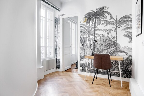 A room with a wooden floor, a desk with a chair, a radiator, and a door leading to another room.
