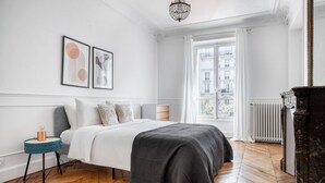 Une chambre à coucher avec un grand lit, une table de chevet, une commode, un lustre et une fenêtre donnant sur des immeubles.