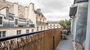 Ein schmaler Balkon mit Holzgeländer, einem Tisch und Stühlen, mit Blick auf eine Reihe von Gebäuden im klassischen europäischen Stil.