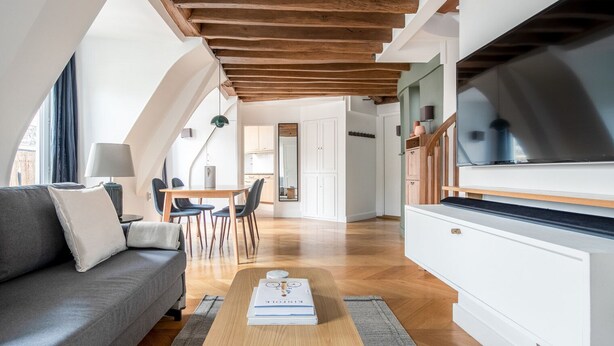 Ein modernes Wohnzimmer mit Sofa, Couchtisch und Fernseher. Es gibt einen Essbereich mit Stühlen und einem Tisch. Das Zimmer hat Holzbalken an der Decke und eine Treppe.
