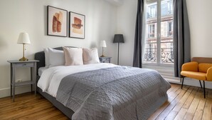 Una camera da letto con un letto, due comodini, una sedia, una finestra con tende e due opere d'arte appese al muro.