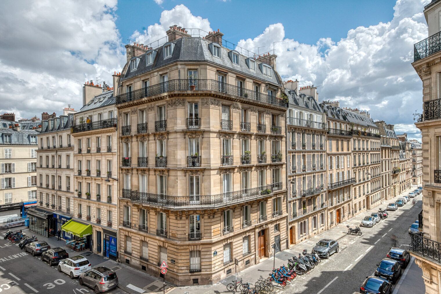 A multi-story building with a classic architectural style, featuring balconies and a street view with parked cars and bicycles.