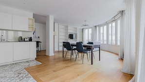 A spacious living area with a dining table, chairs, and a kitchen area with white cabinets and a large window. - Blueground | Sentier-Montorgueil, foodie area (Paris)
