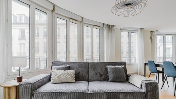 A modern living room with a grey sofa, two pillows, a wooden side table, and a dining area with blue chairs. - Blueground | Sentier-Montorgueil, foodie area (Paris)