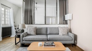 A modern living room with a grey sofa, a wooden coffee table, and a view of a cityscape through large windows. - Blueground | Europe, nr Gare Saint Lazare (Paris)