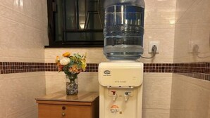 A water dispenser with a large water bottle on top, a wooden cabinet with drawers, and a tiled wall with a window above. - New World Guest House (Kowloon)