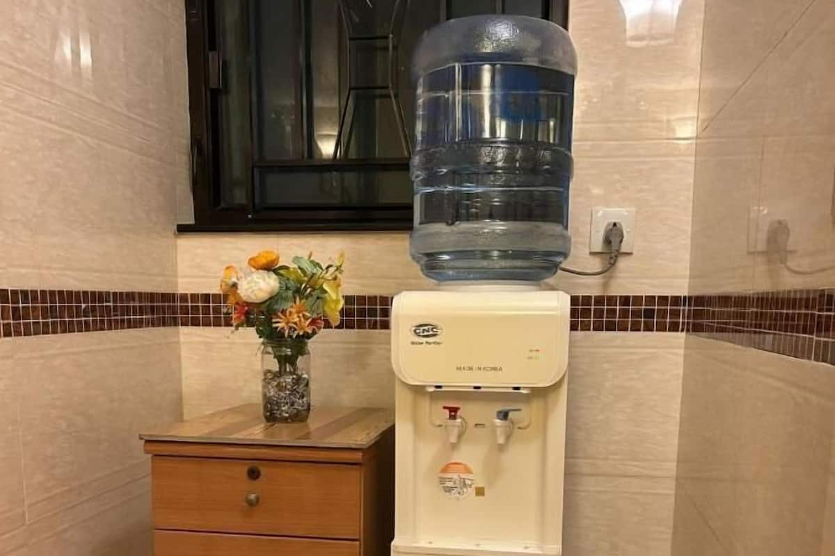 A water dispenser with a large water bottle on top, a wooden cabinet with drawers, and a tiled wall with a window above.