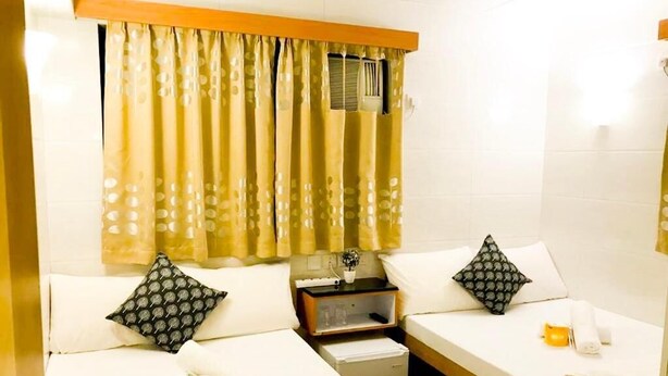 A hotel room with two single beds, a small bedside table with a lamp, a nightstand, and a window with curtains. - New World Guest House (Kowloon)