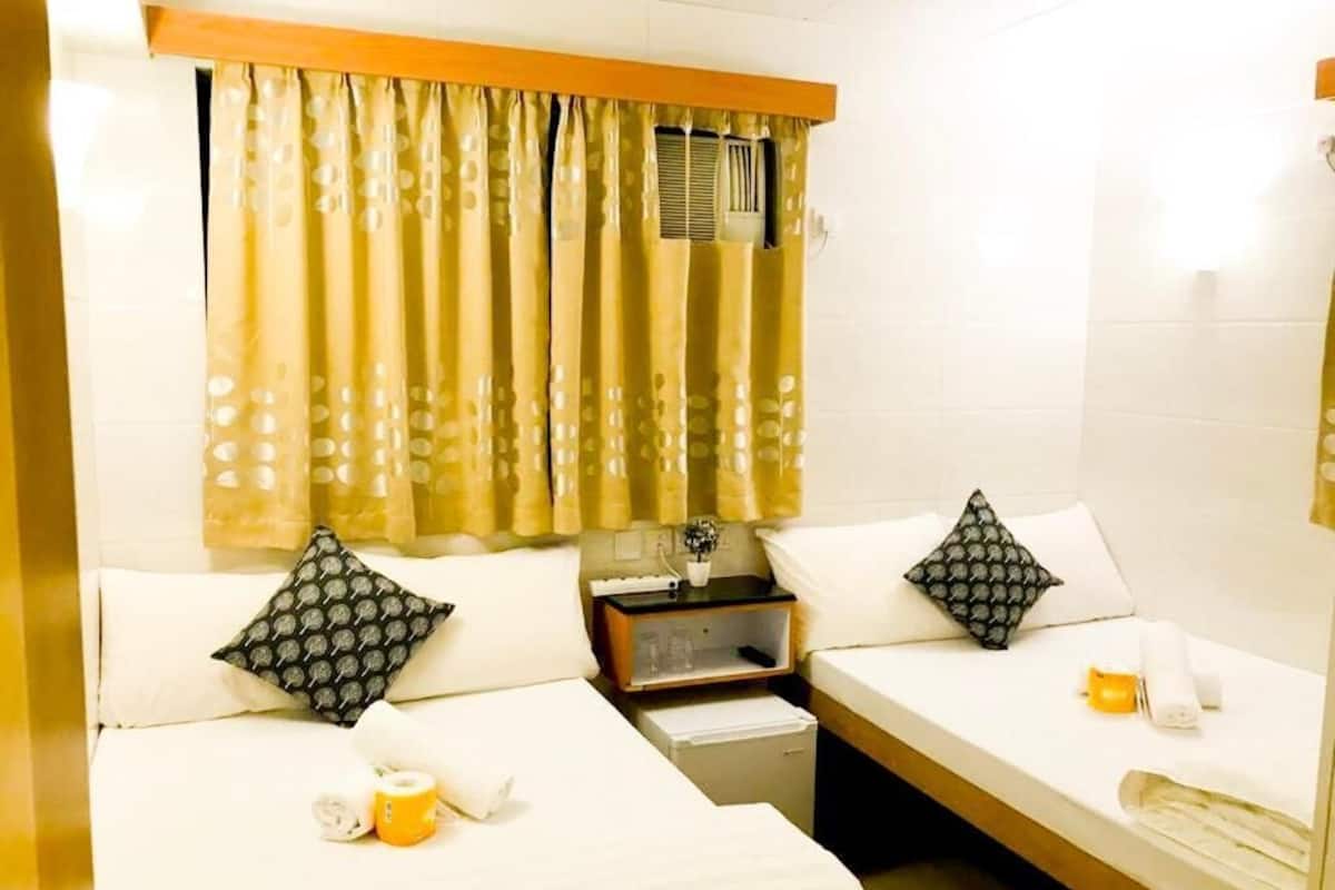 A hotel room with two single beds, a small bedside table with a lamp, a nightstand, and a window with curtains.