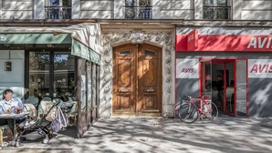 A building with a wooden double door, a cafe with outdoor seating, and a red storefront with the name "AVIS".