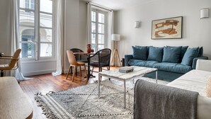 A modern living room with a sofa, coffee table, and chairs. There are large windows with curtains, a floor lamp, and a painting on the wall.