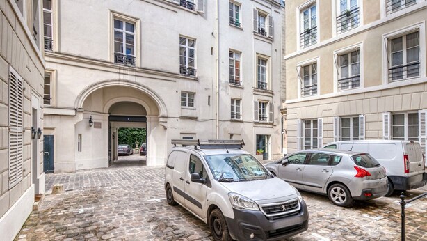 A narrow cobblestone alleyway with a white van parked on the left and a silver car on the right.