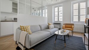 A modern living room with a grey sofa, a coffee table, and a chair. There is a kitchen area with a microwave and a sink. The room has large windows and a chandelier.