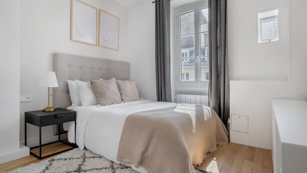 A modern bedroom with a large bed, a bedside table, a lamp, and a view of the city through a window.