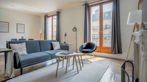 Una sala de estar moderna con un sofá, una mesa redonda de centro y una silla azul. Hay dos ventanas con cortinas, una lámpara de pie y un cuadro en la pared.