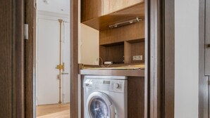 Une petite buanderie équipée d’un lave-linge à chargement frontal, d’étagères en bois et d’une porte donnant sur une autre pièce.