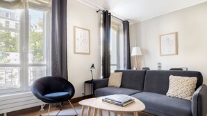 Un salon moderne avec un canapé gris, un fauteuil noir, une petite table ronde sur laquelle sont posés des livres, et deux œuvres d’art encadrées accrochées aux murs.