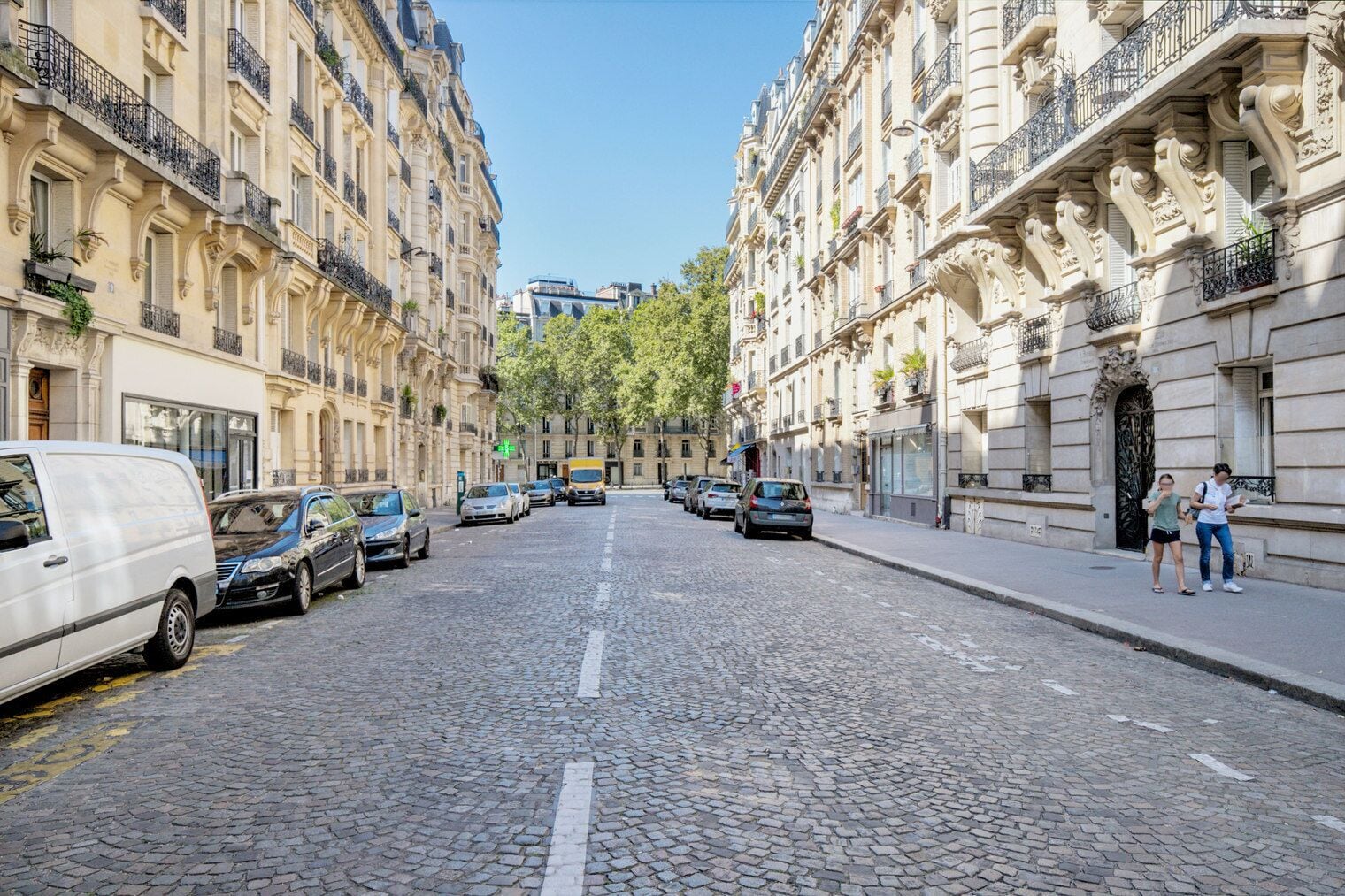 A cobblestone street lined with historic buildings, parked cars, and pedestrians.