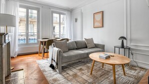 Een ruime woonkamer met een grijze bank, een ronde salontafel, en een eethoek met een tafel en stoelen.
