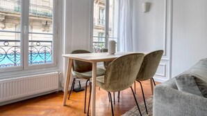 Een kamer met houten vloer, een tafel met twee stoelen, een radiator en een raam met uitzicht op gebouwen en een balkon.