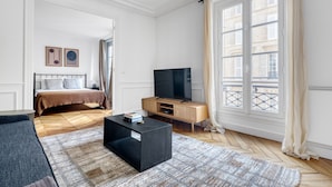 Une chambre moderne avec un lit, une télévision, une commode en bois et une grande fenêtre avec des rideaux.