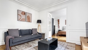 Un salon moderne avec un canapé gris, une table basse noire et un tapis à motifs. On trouve une bibliothèque et un lit dans la pièce adjacente.