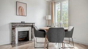 A modern dining area with a marble fireplace, two grey upholstered chairs, a wooden table, and a large window with a view of trees. - Blueground | Beaugrenelle, nr Seine & mall (Paris)
