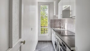 A narrow kitchen with white cabinets, a stainless steel sink, and a window with a view of trees. - Blueground | Beaugrenelle, nr Seine & mall (Paris)