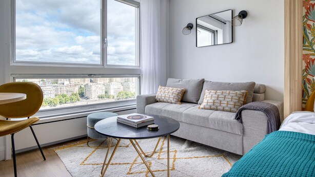 Une chambre moderne avec un canapé gris, une table basse ronde, un fauteuil jaune et une grande fenêtre donnant sur la ville.