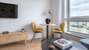 Un salon moderne avec un meuble en bois, deux fauteuils jaunes, une table ronde sur laquelle repose un livre, et une vue sur un paysage urbain par la fenêtre.