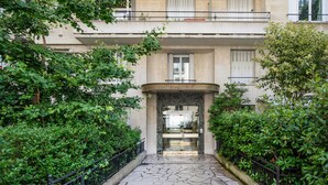 A stone-paved pathway leading to a building entrance with a glass door, surrounded by lush greenery and a metal railing. - Blueground | La Muette, elev & balcony, nr mall (Paris)