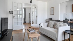 A modern living room with a white sofa, two side tables, a bookshelf, and a dining area with a table and chairs. - Blueground | La Muette, elev & balcony, nr mall (Paris)
