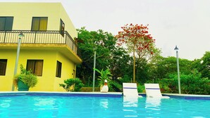 Una piscina con un edificio amarillo al fondo, rodeada de follaje y un árbol con flores rojas.
