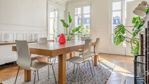 Une salle à manger avec une table en bois, six chaises, un vase rouge et de grandes plantes vertes.