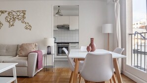 En moderne stue med sofa, spisebord med stole, et køkken i baggrunden og udsigt over byen gennem store vinduer.