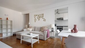 En moderne stue med sofa, sofabord og et spiseområde med bord og stole. Et køkken kan ses gennem en åben dør.