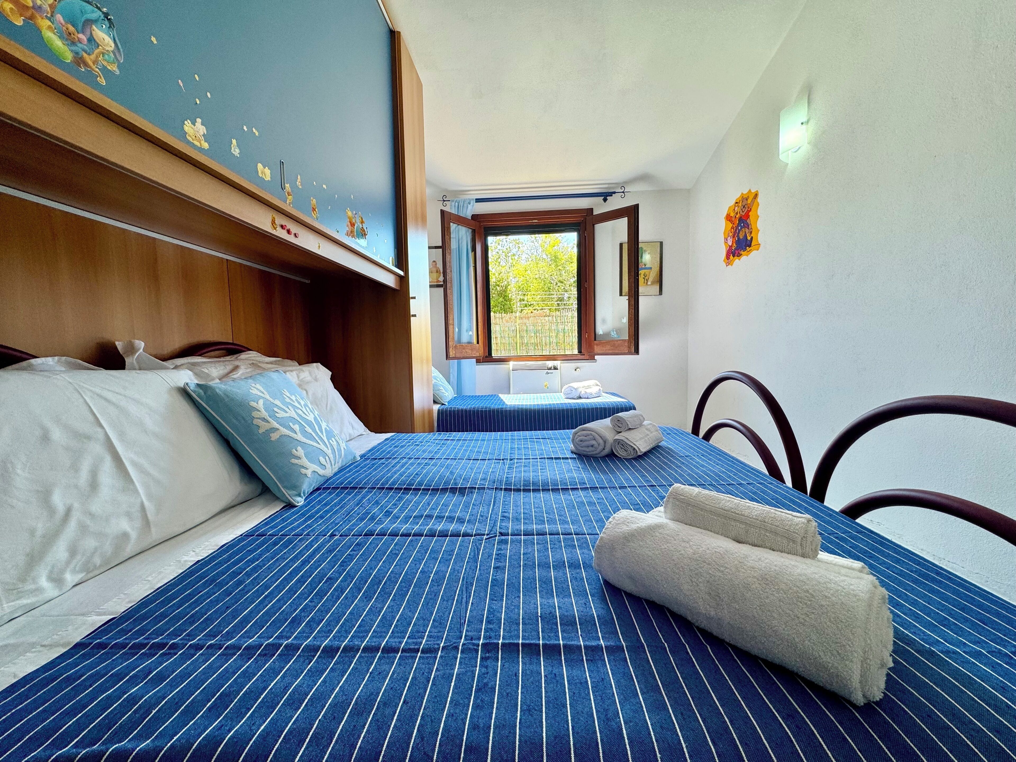 Une chambre d’hôtel avec un grand lit, une fenêtre, une tête de lit en bois et un mur orné d’une fresque.