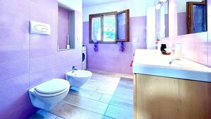 A bathroom with purple tiles, a toilet, a bidet, a sink, and a window with a view of greenery.