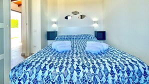 Une chambre à coucher comprenant un lit, deux tables de chevet, une tête de lit agrémentée d’un miroir et une porte donnant sur un espace extérieur lumineux.
